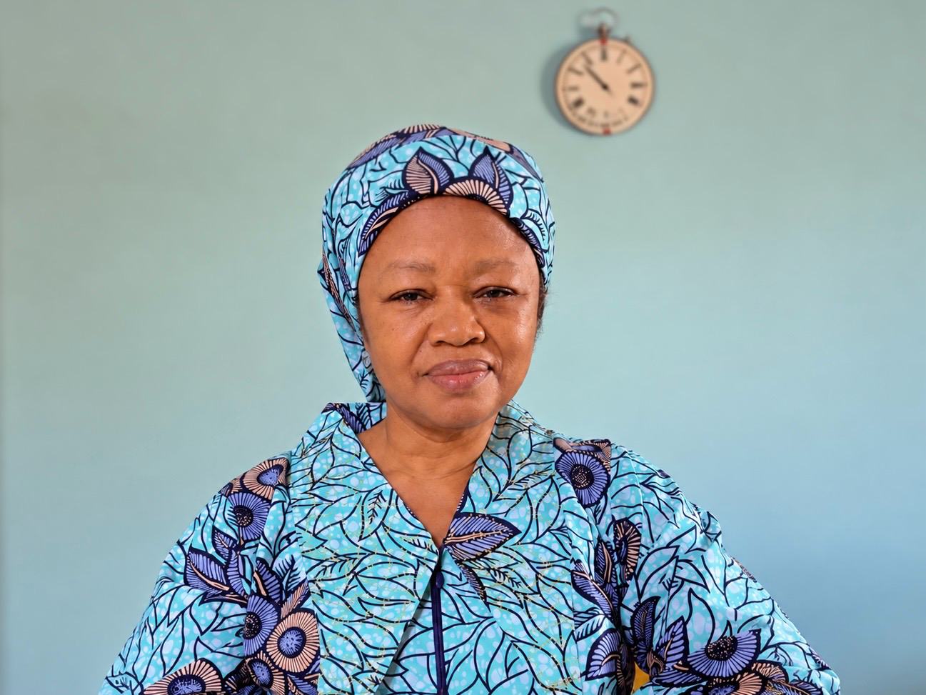 Mrs Egbo in a turquoise and violet floral print and headwrap, a clock on the wall behind her